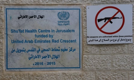 Signs at the clinic – one shows a weapon in a red circle with a line through it with text underneath in Arabic and English reading ‘no weapons are authorised within these premises’.