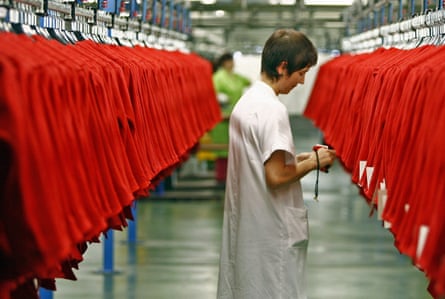 A woman stands on a factory floor between two long racks of hanging red jackets.