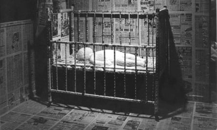 Black and white photo from 1976 showing baby made from cake in a cot against background of walls papered with newspaper