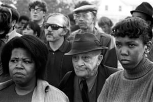 Black and white photo. Two black women and a white man with a hat frown in the front row of a crowd