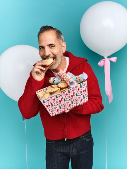 Yotam Ottolenghi holding a Christmas paper-wrapped box of mince pies and crackers in one hand, eating a mince pie with the other