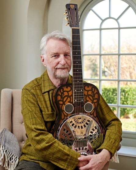He holds a guitar in his arms, while posing for a portrait
