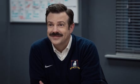Jason Sudeikis, dengan kumis dan senyuman, mencondongkan tubuh ke depan