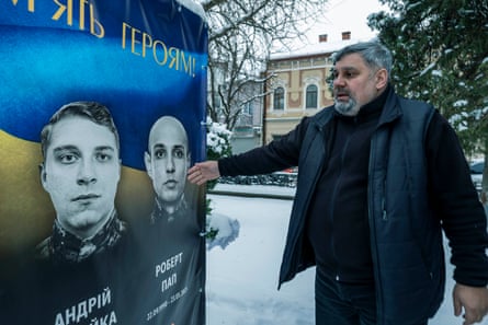 A man standing on a snowy street gestures to one of two images of young men’s faces on a poster