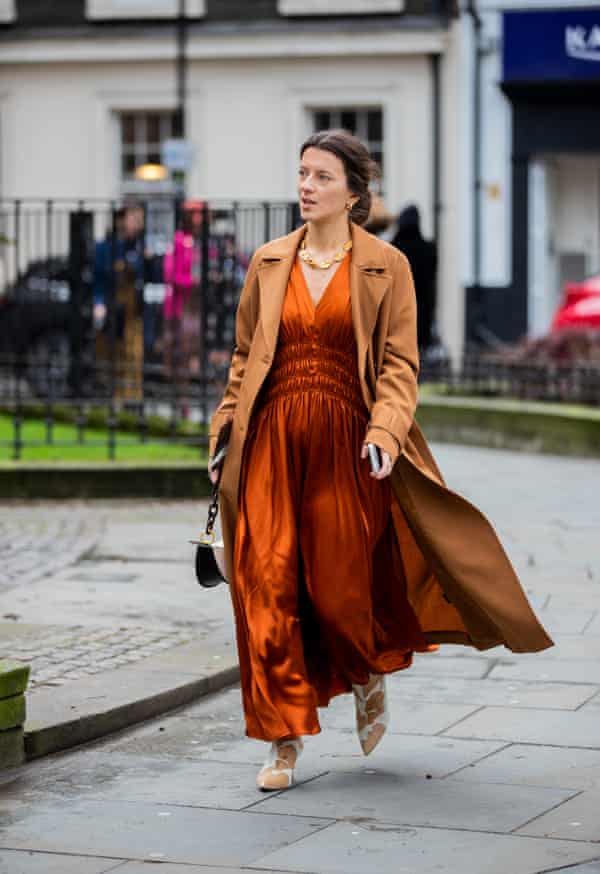 A woman wearing an orange silk dress and a brown coat walking along a paved path outside Erdem during London fashion week