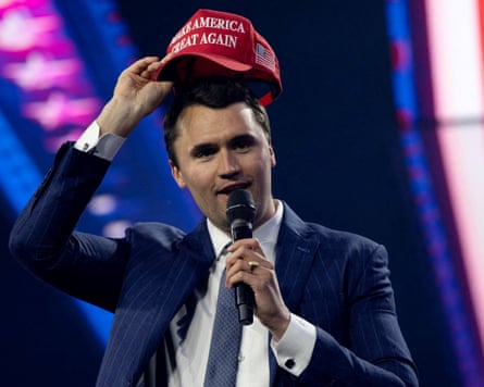 Kirk holds a microphone while he adjusts the red Maga cap he is wearing