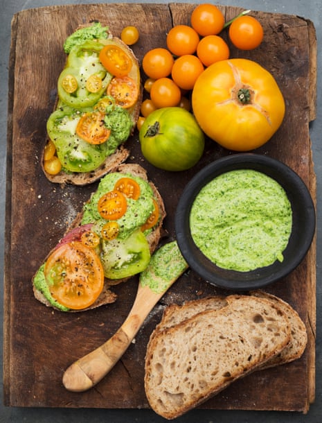 A wooden board with pieces of brown bread topped with large slices of tomato and green pesto, a bowl of pesto and tomatoes