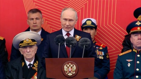 Vladimir Putin is photographed from the front standing at a podium, speaking into microphones, in Moscow's Red Square. Behind him are several uniformed Russian military service members, standing against a red backdrop.