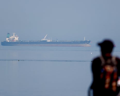 A large oil tanker is visible in calm waters, with a blurred figure watching in the foreground