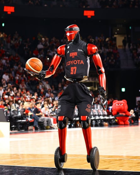 CUE7, a red robot with wheels for feet and nets for hands, holds a basketball on a court
