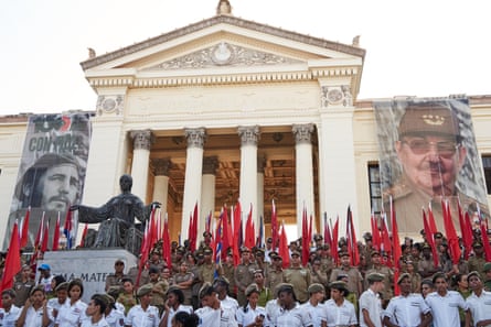 A large crowd of people in military uniforms stand in front of a neo-classical building with columns decorated with banners of Fidel and Raúl Castro and red flags