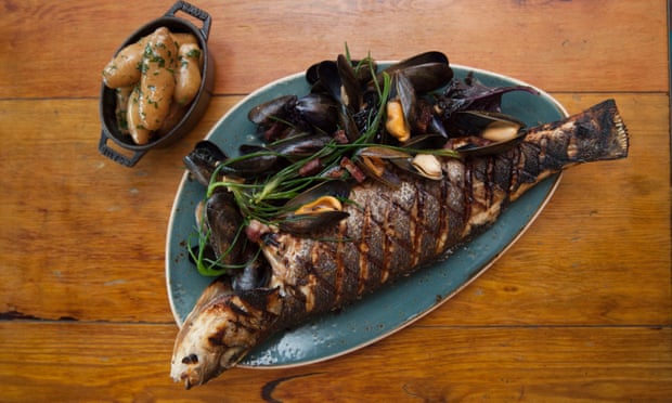 A whole crispy skinned sea bass with the head and tail hanging over the ends of the blue plate, mussels by the side and a small bowl of buttered new potatoes