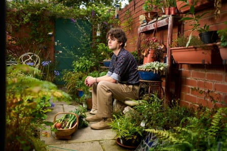 Samuel Rowe sits against a brick wall in his garden amid pots of flowers and herbs and beds with feathery ferns; he is 35 with dark wavy hair and wears light tan trousers and a dark blue patterned shirt. A trug filled with vegetables is by his feet and plants are on shelves on the wall as well as on the paving.