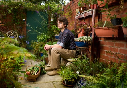Samuel Rowe sits against a brick wall in his garden amid pots of flowers and herbs and beds with feathery ferns; he is 35 with dark wavy hair and wears light tan trousers and a dark blue patterned shirt. A trug filled with vegetables is by his feet and plants are on shelves on the wall as well as on the paving.
