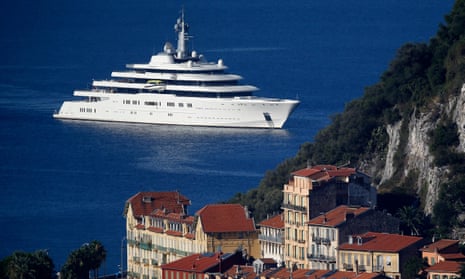 A very large, maybe six-story-high white yacht, in deeply blue water off a steep, rocky, forested coast that also has narrow, red-roofed buildings.