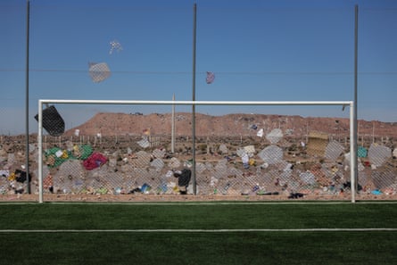 A football goal stands on artificial turf next to a fence. The fence is plastered with bags and rubbish and behind it can be seen the mounds of the tip