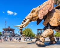 A giant elephant puppet made of wood walks through a pedestrian square in the sun