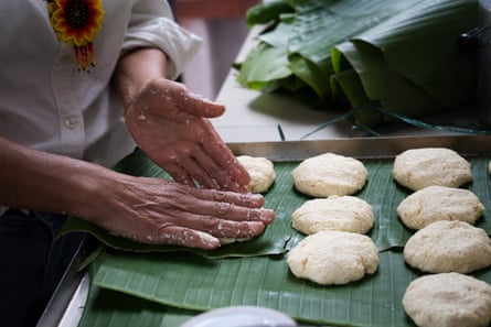 A woman makes shapes corn dough into flat, round shapes laid out on large green leaves on a work bench