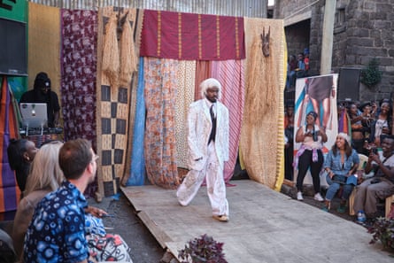 A man wearing a blonde wig and a white oversized suit covered in silver applique strides out on the runway.