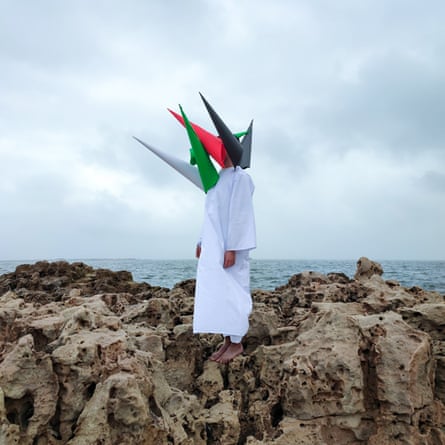A man wearing a white robe, his face covered with several cones in the different colours of the Sudanese flag, stands on a rocky outcrop by the sea.