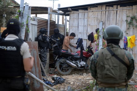 Masked paramilitary police and soldiers stand in the yard of a shack as two young men and a woman stand by a motorbike