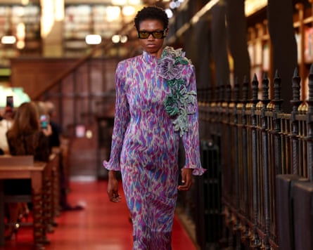 A model walks down the catwalk in a purple dress and dark sunglasses as spectators holding up phones seated in pews