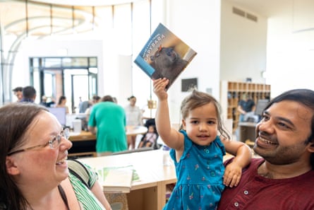 A man carries a little girl who is holding a book in the air, next to a smiling woman in a public library.