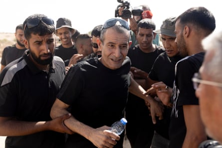 A man, surrounded by and being held by other men, walks forward carrying a bottle of water