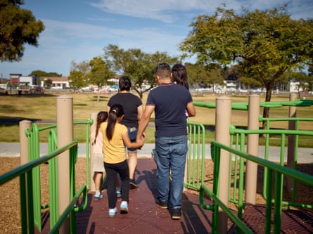 a man holding his daughter in one arm and the other daughter by the hand walks through the park with his wife and other daughter