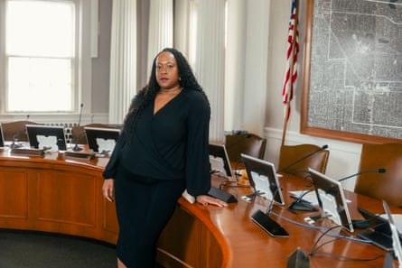 A Black woman with long black curly hair wearing a black dress stands in front of a brown desk used by the city council in Shaker Heights, Ohio. On the right, there is an American flag and a framed map on the wall.