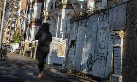 A young woman walks down a British street, with graffiti adorning the wall nearest to her qhiqhhiqhiruinv