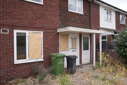 A brick house with a white front door and small porch has its windows and door boarded up after glass was smashed