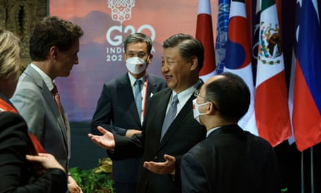 Justin Trudeau and Xi Jinping, flanked by two officials, face each other in greeting next to several flags hanging from poles