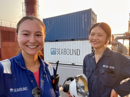 Two young women smile standing in front of a stack of shipping containers.