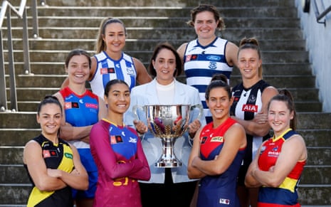 A woman holding a large silver trophy is flanked by four women on each side wearing different football jerseys