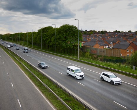 A motorway with vehicles traveling alongside trees and brick housing estates in Rochdale under a cloudy sky