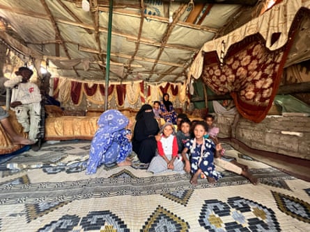 Two women and a group of children sit on a rug on the floor of a tent in a refugee camp