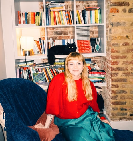 Megan Nolan at her home, sitting on a dark blue upholstered chair, a bookcase full of books - and a black cat - behind her