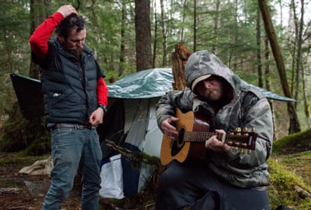 Two men in the woods next to a tent, one playing guitar.