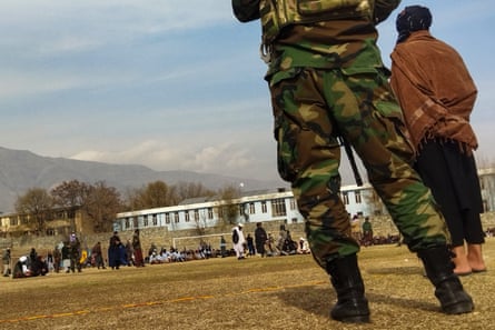 A soldier in camouflage fatigues and a Talib wearing traditional Afghan dress seen from behind at a stadium with people sitting down and being guarded in the background