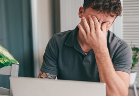 A young man rubs his forehead anxiously in front of a laptop