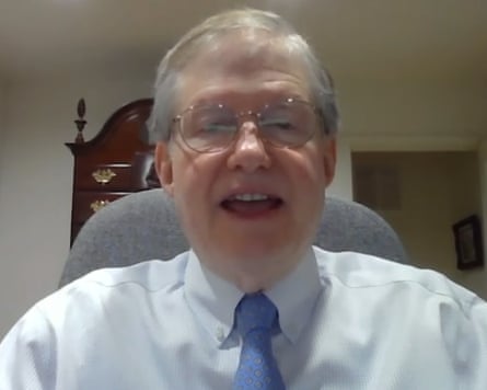 A screengrab showing him sitting in a chair on a video call. He wears a light-blue shirt and a darker blue tie