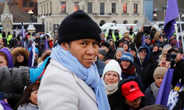 Woman wearing a black beanie, white coat and blue scarf looking into the camera. A crowd can be seen in the background.