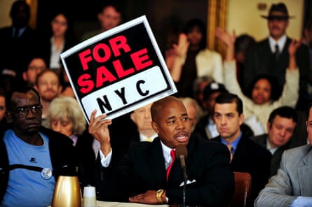 a man holds up a red, black and white sign that says ‘for sale NYC’, as a crowd of people stand behind him and look on