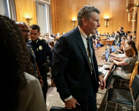 Police officers walk behind an attorney dressed in a suit as he leaves a large wood-panelled room