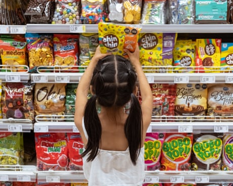 A young Latina girl seen from behind taking a packet of sweets from among several shelves of sweets and snacks