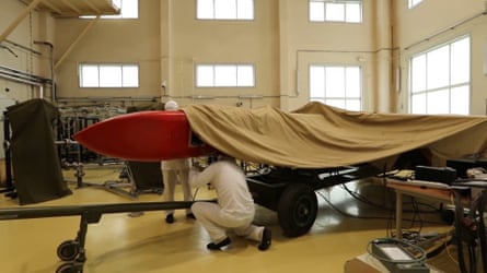 it is red and its nose is pointing out from under a canvas sheet while two men in white overalls appear to be securing it on a trailer. They are within a high-roofed, white-painted factory-style building with tall windows.
