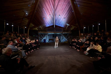 Ian McKellen stands in the centre of the performance space with people in tiered seating to either side and the front. Behind him is scaffolding and a raised platform, and above is an illuminated wooden ceiling, arched like the hull of a boat.