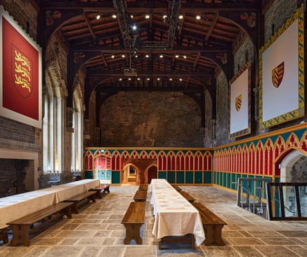 A high-ceilinged medieval dining hall with long tables and benches, heraldic wall hangings and red and green decorated panelling