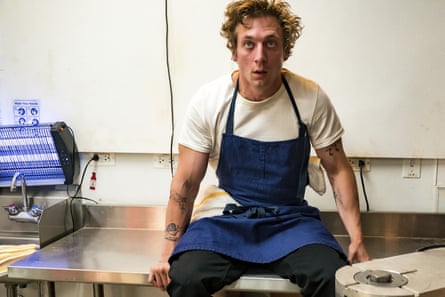 a man in a white T-shirt and blue apron sits in a restaurant kitchen